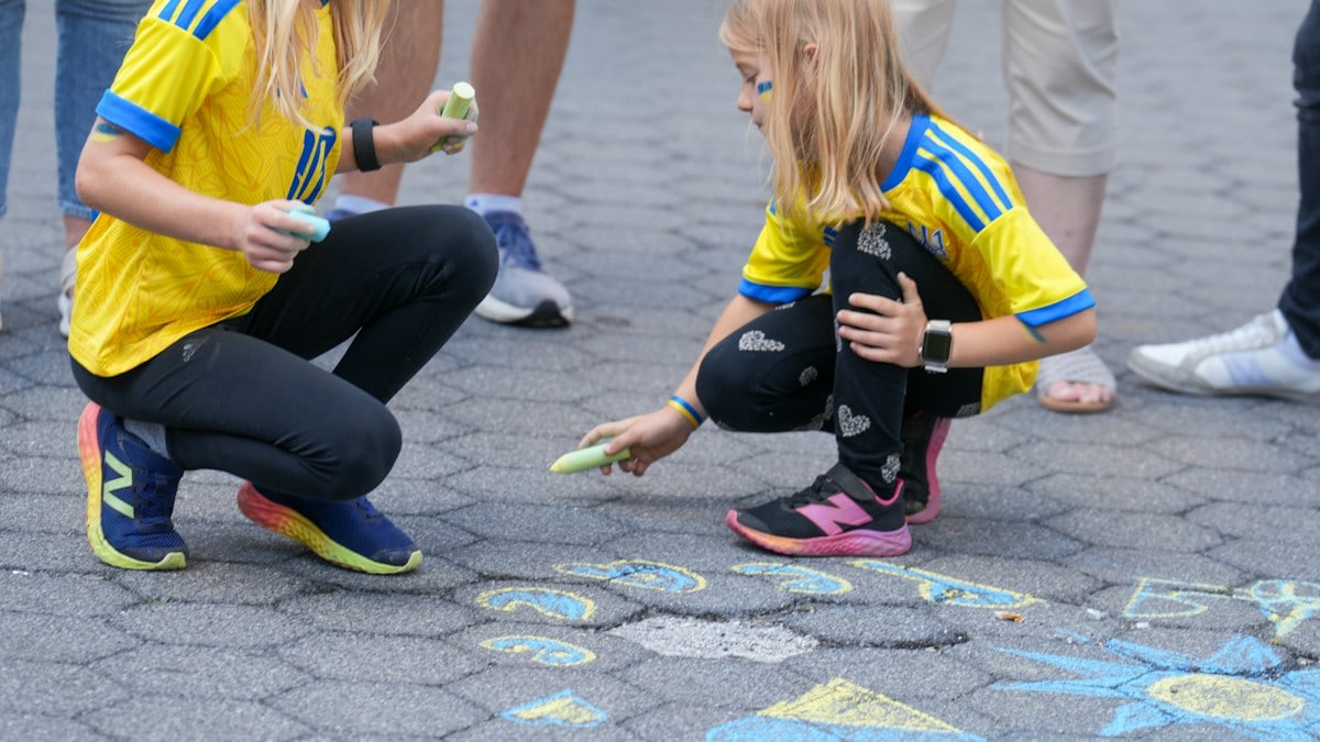 A smiling young woman with a blonde side braid stands outdoors at a street gathering, wearing a white Ukrainian embroidered vyshyvanka blouse and pleated light blue skirt. She carries a small taupe crossbody bag holding miniature American and Ukrainian flags, with people in embroidered shirts and a yellow flag blurred behind her. Scenes from a Ukrainian anti-war rally in downtown Manhattan, where demonstrators gathered with flags and signs to protest the war and show solidarity with Ukraine.